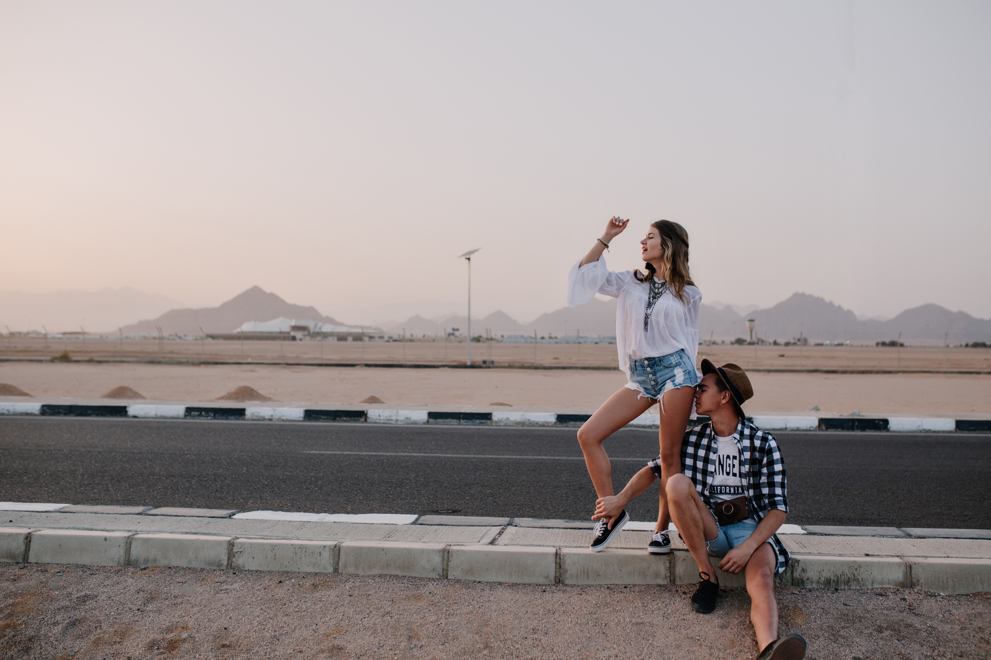 امرأة نحيفة مرحة ترقص بشكل مضحك بينما صديقها المتعب يستريح على الطريق الجبلي صورة امرأة شابة رائعة رجل يسافر حول البلاد في انتظار ركوب الطريق السريع